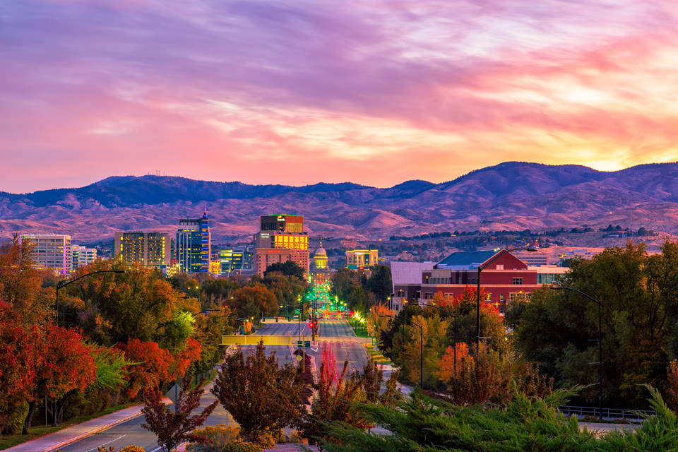 The Idaho State Capitol at sunset, seen down a tree-lined street with downtown Boise and the mountains behind it