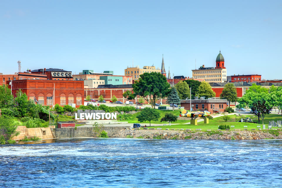 A "LEWISTON" sign on the riverbank with the historic mill buildings and Great Falls behind it in Lewiston, Maine