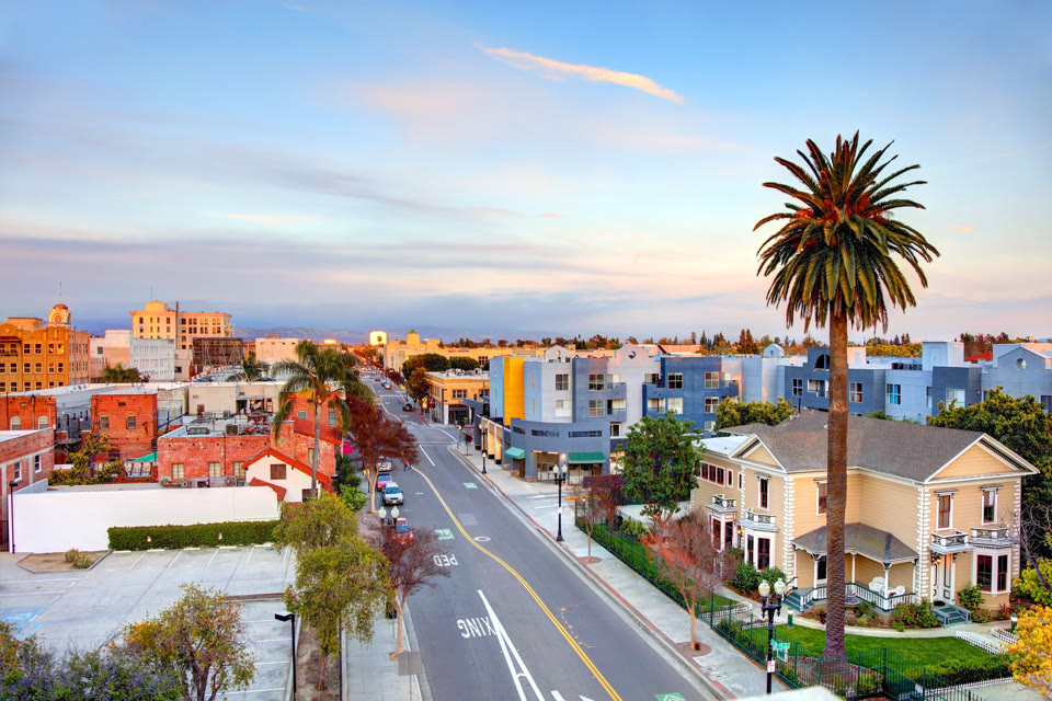 A historic downtown Santa Ana, California street lined with Victorian homes and a palm tree at dusk, with the city's downtown skyline visible in the distance